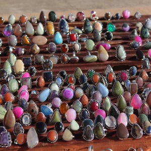 Puede incluir: Una gran colección de anillos con diversas piedras preciosas pulidas en formas de lágrima, ovaladas y redondas. Los anillos están montados en bandas de metal y dispuestos en filas sobre un expositor de madera oscura, mostrando gran variedad.