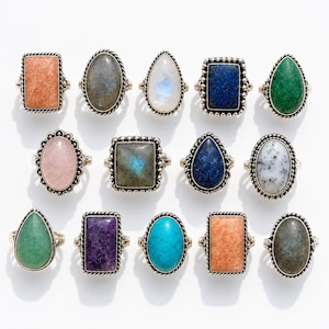 May include: A collection of fourteen rings featuring various gemstones in oval, rectangular, and teardrop shapes. Each ring has a silver-toned metal band with a twisted rope-style border surrounding the stone, showcasing diverse colours and textures.