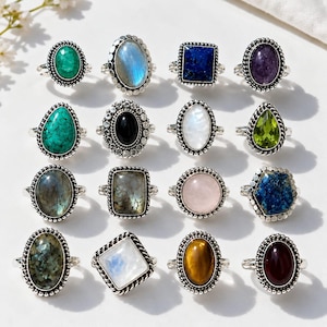 May include: A collection of sixteen silver-toned rings arranged in a four-by-four grid. Each ring features a unique gemstone, including turquoise, moonstone, lapis lazuli, amethyst, peridot, rose quartz, tiger's eye, and various other mineral stones.