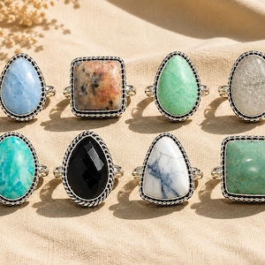 Puede incluir: Una colección de ocho anillos con diversas piedras preciosas engastadas en monturas de metal plateado con bordes de cuentas. Las piedras son azul claro, beige moteado, verde claro, gris, turquesa, negro, blanco con vetas grises y verde sólido.