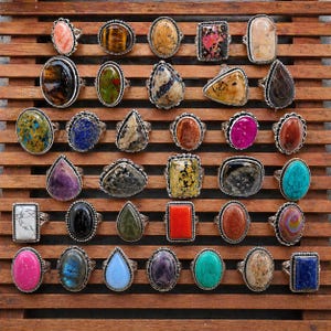 May include: A collection of thirty rings featuring various polished gemstones in diverse shapes and colors, including oval, pear, rectangular, and round settings. Each ring is crafted with a silver-toned metal band and bezel, showcasing unique natural stone patterns and vibrant hues.