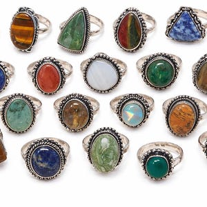 May include: A collection of silver rings with various gemstone settings. The rings feature stones in shades of purple, brown, green, blue, and orange. The gemstones are oval, triangular, and square-shaped. The rings are displayed on a white surface.