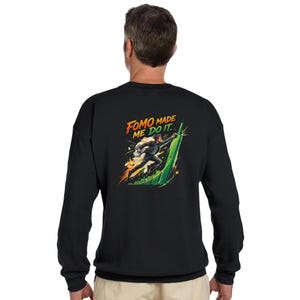 Puede incluir: Sudadera negra con un gráfico de una persona corriendo por una estructura verde, con el texto "FOMO MADE ME DO IT." El diseño presenta acentos naranjas, amarillos y blancos. La sudadera es de cuello redondo y manga larga.