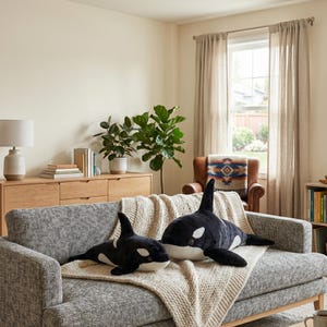 May include: Two plush orca whale stuffed animals, one large and one small, rest on a gray sofa with a cream-colored blanket. The whales are black and white, with detailed features. The scene is set in a well-lit living room.