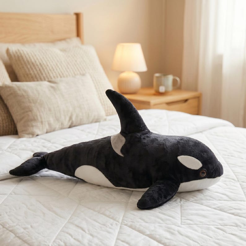 May include: A plush, black and white orca whale stuffed animal with a dorsal fin, pectoral fins, and a white belly. The toy is lying on a white quilted bedspread. The orca has white patches around its eyes.