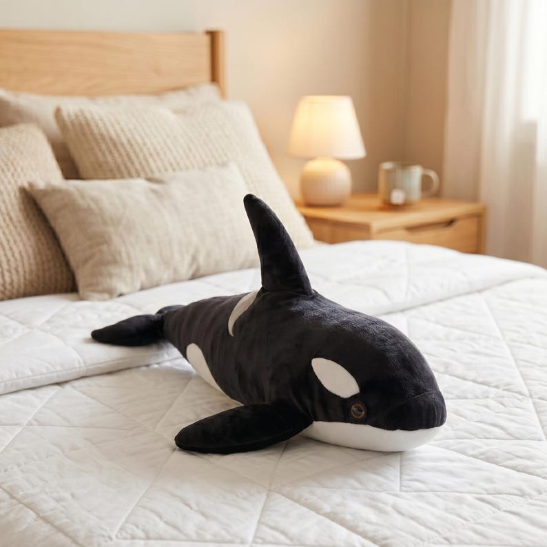 May include: A plush orca whale toy in black and white, with a dorsal fin and flippers. The stuffed animal is lying on a white quilted bedspread. The toy has white patches around the eyes and on the sides.