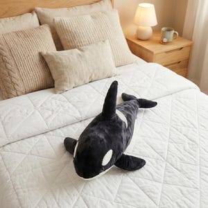 May include: A plush, black and white orca whale stuffed animal rests on a white quilted bedspread. The orca has a distinctive black dorsal fin, white patches, and flippers. The bed is made up with neutral-toned pillows and a wooden nightstand.