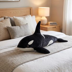 May include: A plush, black and white orca whale stuffed animal with a soft, velvety texture. The toy features white markings around the eyes and belly, and is lying on a bed with neutral-toned bedding. The orca measures approximately 36 inches long.