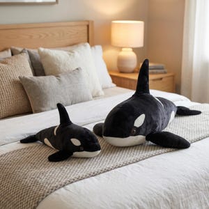 May include: Two plush orca whale stuffed animals in black and white, one large and one small, are displayed on a bed with neutral-toned bedding. The whales have white patches and fins, and are positioned on a bedspread with a neutral-colored runner.