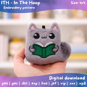 Puede incluir: Un gato de peluche gris sosteniendo un libro verde, un patrón de bordado en el bastidor. El gato tiene grandes ojos negros y el interior de las orejas rosas. El texto "ITH - In The Hoop Embroidery pattern" está en la parte superior. Tamaño 10.16 x 10.16 cm. Descarga digital.