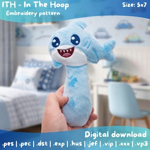 以下が含まれることがあります： 手で持たれた、刺繍が施されたディテールのライトブルーのぬいぐるみサメのおもちゃ。サメは白いお腹、赤い口、大きな青い目をしています。「ITH - In The Hoop Embroidery pattern」の文字が見えます。サイズは12.7 x 17.8 cmです。デジタルダウンロード。