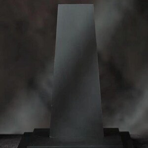 May include: A black, rectangular display stand with a trapezoidal top. The stand has a tiered base and a dark, matte finish. The text on the base reads "Complimentary Base".
