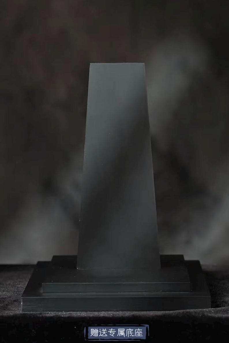 May include: A black, rectangular display stand with a trapezoidal top. The stand has a tiered base and a dark, matte finish. The text on the base reads "Complimentary Base".