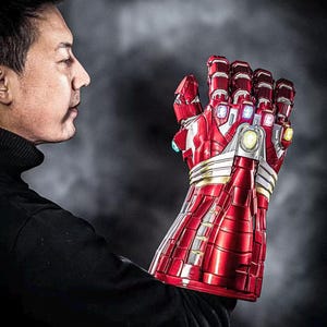 May include: A red and silver Iron Man gauntlet with six colored gems. The gauntlet is highly detailed with a metallic finish and is held up against a dark background. The gauntlet is a replica of the one used by Iron Man.