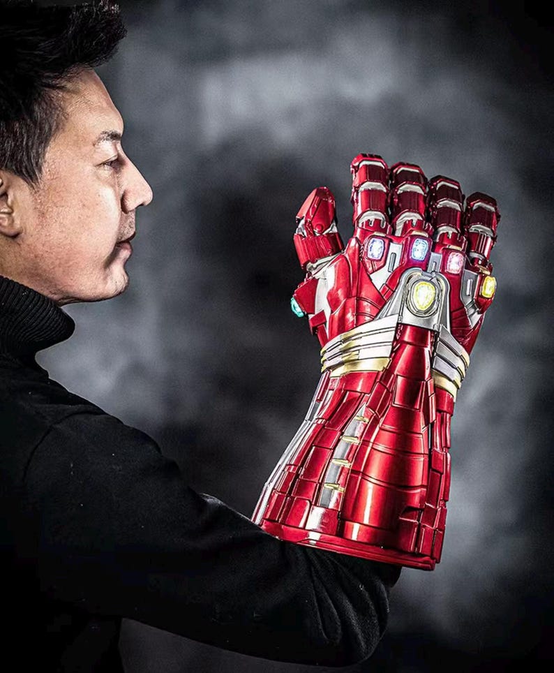 May include: A red and silver Iron Man gauntlet with six colored gems. The gauntlet is highly detailed with a metallic finish and is held up against a dark background. The gauntlet is a replica of the one used by Iron Man.