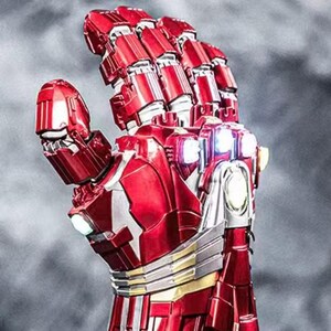 May include: A detailed, red and silver robotic hand with articulated fingers. The metallic gauntlet features intricate paneling, gold accents, and glowing lights, set against a blurred gray background. The design is reminiscent of a superhero's armor.