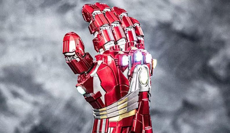May include: A detailed, red and silver robotic hand with articulated fingers. The metallic gauntlet features intricate paneling, gold accents, and glowing lights, set against a blurred gray background. The design is reminiscent of a superhero's armor.