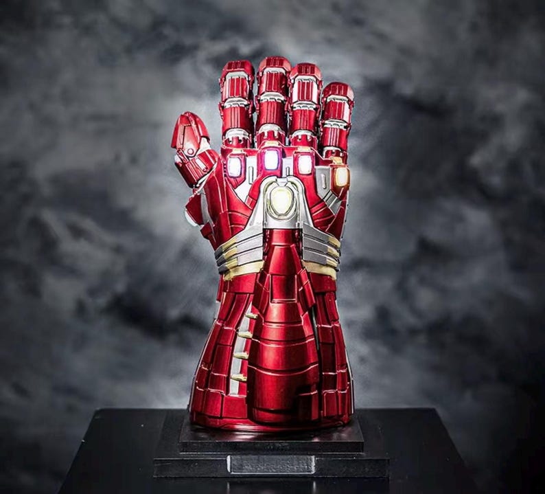 May include: A red and silver metallic gauntlet with a detailed design, featuring articulated fingers and glowing accents. The gauntlet is displayed on a black base, set against a dark, smoky background. A collectible item.