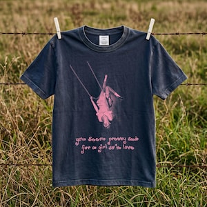 Könnte beinhalten: Ein dunkel anthrazitfarbenes Kurzarm-T-Shirt mit einer rosa Grafik, die eine Person zeigt, die kopfüber auf einer Schaukel hängt. Unter der Grafik steht der Text "you seem pretty sad for a girl so in love" in einer rosa, geschwungenen Schrift.