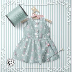 Peut inclure: Une robe sans manches bleu clair avec un motif de lapin blanc et rose. La robe a une fermeture boutonn&eacute;e blanche et est suspendue &agrave; un cintre en bois. La robe est sur un fond ray&eacute;.