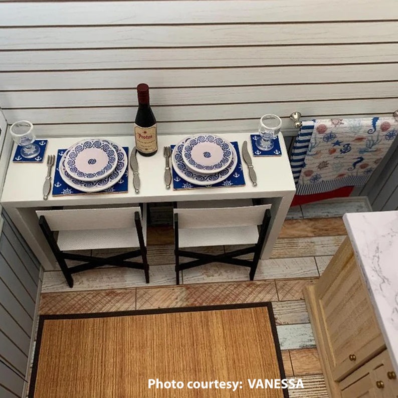 May include: A white table with two place settings, each with a blue and white patterned plate, a blue and white patterned coaster, a fork, a knife, and a glass. A bottle of wine is on the table between the place settings. Two white chairs with black legs are in front of the table. The table is in a room with wood floors and white walls.
