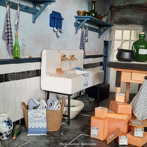 May include: A miniature kitchen scene with a white porcelain sink with two faucets, a wicker basket filled with blue and white towels, and a wooden table with stacked boxes. The walls are painted blue and white, and there are various kitchen utensils hanging on the wall.