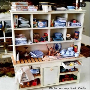 May include: A white miniature hutch with a wooden countertop and open shelves. The hutch is filled with miniature kitchen items, including dishes, towels, and food items. The hutch has two drawers and a cabinet door.