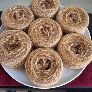Peut inclure: Sept pelotes de laine aux tons bruns, beiges et crème sont disposées sur une assiette blanche. La laine est enroulée en boules rondes, avec un aspect texturé et multicolore, idéal pour les projets de tricot ou de crochet.