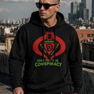 Puede incluir: Sudadera con capucha negra con un gráfico rojo y verde. El gráfico incluye las palabras "DISSBEY", "BE A DISSIDENT", "DESTROY SYSTEM" y "JOIN TO THE CONSPIRACY". La sudadera tiene un bolsillo delantero y una capucha con cordón.