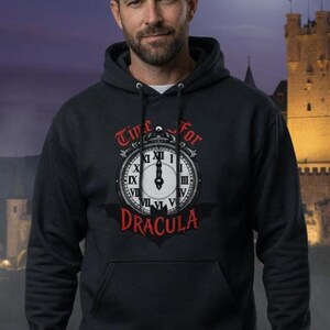 Puede incluir: Sudadera con capucha gris oscuro con un diseño de reloj que dice "Time for Dracula". El gráfico del reloj es blanco con números romanos negros y una silueta de murciélago. La sudadera tiene un bolsillo delantero y una capucha con cordón.