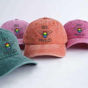 Può includere: Una collezione di cappellini da baseball in vari colori, tra cui verde acqua, rosa e corallo. Ogni cappellino presenta la scritta ricamata "BEE PROUD" con un'ape arcobaleno. I cappellini hanno un aspetto vintage.