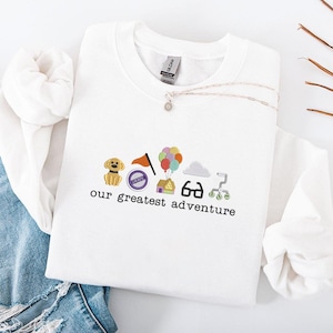 Puede incluir: Sudadera blanca de cuello redondo con elementos gráficos coloridos bordados, que incluyen un perro, una bandera, una casa, globos y el texto "our greatest adventure". Un collar plateado con un colgante está drapeado sobre la sudadera.