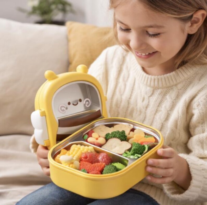 Pu&ograve; includere: Un portapranzo giallo e bianco con un design a forma di ape dei cartoni animati, aperto per rivelare un pasto di verdure e frutta. Il portapranzo ha un interno in acciaio inossidabile ed &egrave; diviso in scomparti. Il bambino che tiene il portapranzo indossa un maglione color crema.