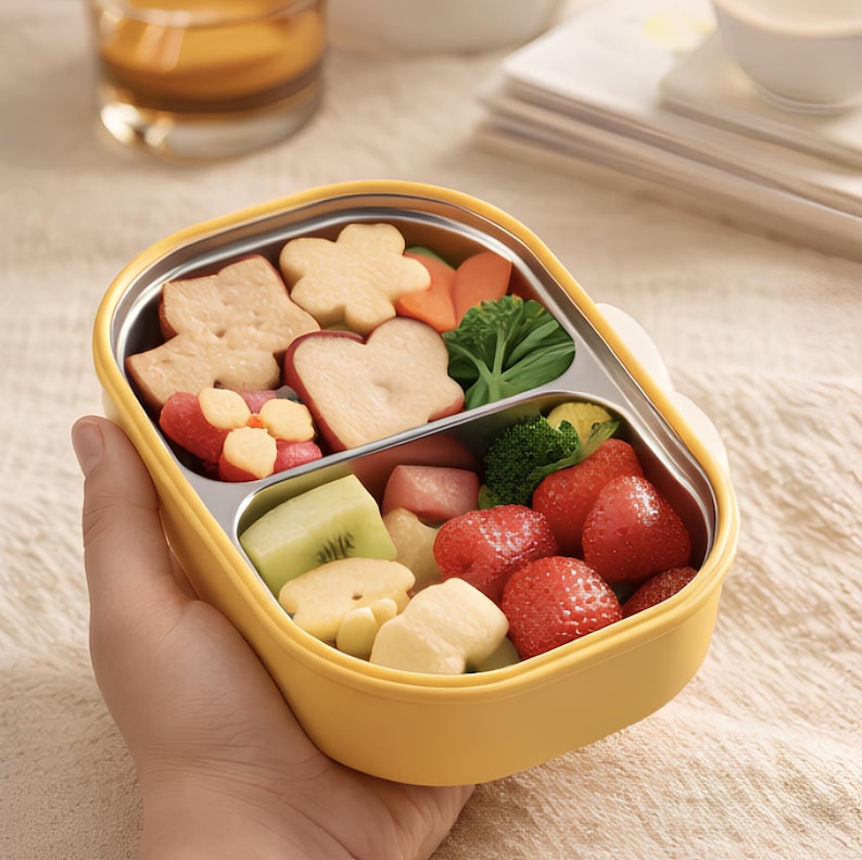 Pu&ograve; includere: Un bento box giallo in acciaio inossidabile pieno di una variet&agrave; di frutta e verdura colorata tagliata. La scatola contiene fragole, kiwi, broccoli e cracker a forma di fiore. La scatola &egrave; tenuta in mano.