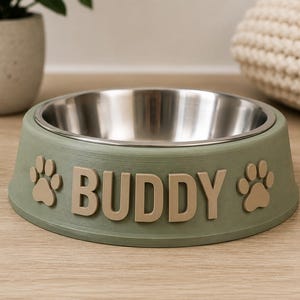 Puede incluir: Un cuenco para mascotas de color verde salvia con un inserto interior de acero inoxidable. El exterior presenta el nombre BUDDY en letras beige en relieve, flanqueado por dos iconos de huellas beige a cada lado del nombre. Tiene una base ancha.