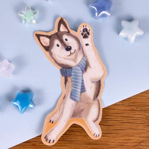 Könnte beinhalten: Ein gestanzter Aufkleber, der einen braun-weißen Husky mit einem blau-weiß gestreiften Schal zeigt, der winkend mit einer erhobenen Pfote dargestellt ist. Die Illustration hat einen hellbraunen Rand und ist von kleinen, durchscheinenden sternförmigen Perlen umgeben.