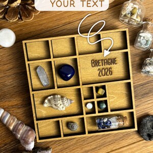 May include: A square wooden display box with multiple compartments containing various natural stones, seashells, and small glass bottles with cork stoppers. One compartment is engraved with the text BRETAGNE 2026. The box is arranged on a wooden surface.
