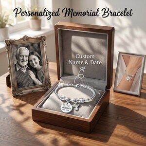 K&ouml;nnte beinhalten: Ein personalisiertes Gedenkarmband mit einem runden Anh&auml;nger mit Gravur von Namen und Daten, pr&auml;sentiert in einer Holzbox. Das Armband hat Charms mit dem Wort "LOVE" und einem kleinen Engel. Das Bild zeigt auch ein gerahmtes Foto.