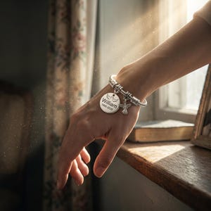 K&ouml;nnte beinhalten: Ein silbernes Charm-Armband mit mehreren Anh&auml;ngern. Ein Anh&auml;nger ist ein runder Anh&auml;nger mit dem Namen "William" und den Daten "1955-2026". Weitere Anh&auml;nger sind ein Engel und ein Paar Fl&uuml;gel. Das Armband wird am Handgelenk getragen.