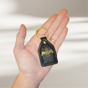 May include: A black leather keychain with a gold-colored eagle design. The keychain has a gold-colored ring and a black leather strap.