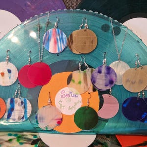 Può includere: Orecchini fatti a mano con vari disegni colorati, tra cui motivi astratti e colori uniti. Gli orecchini sono esposti su un supporto trasparente ad arco, con dischi in vinile sullo sfondo.