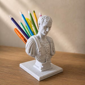 May include: A white pen holder shaped like a bust of a Roman emperor, holding several pens and pencils. The pens and pencils are various colors, including orange, blue, green, and yellow. The pen holder is on a wooden surface.