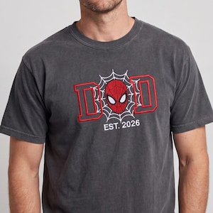 K&ouml;nnte beinhalten: Ein anthrazitfarbenes T-Shirt mit einem rot-wei&szlig;en Stickdesign. Das Design zeigt das Wort "DAD" mit einer Spider-Man-Maske in der Mitte und dem Text "EST. 2026" darunter. Das T-Shirt ist kurz&auml;rmlig.