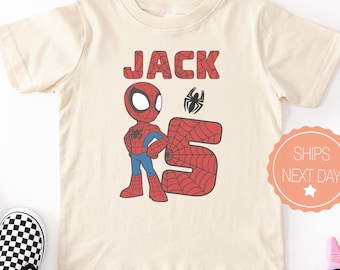May include: A cream-colored short-sleeve t-shirt featuring a graphic of a cartoon superhero, the name JACK in red web-patterned letters, and a large red web-patterned number 5. A small black spider icon is positioned above the number 5.