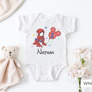 White baby onesie with custom Spiderman birthday design, personalized name Nathan and age 1, colorful stars, styled with teddy bear and flowers