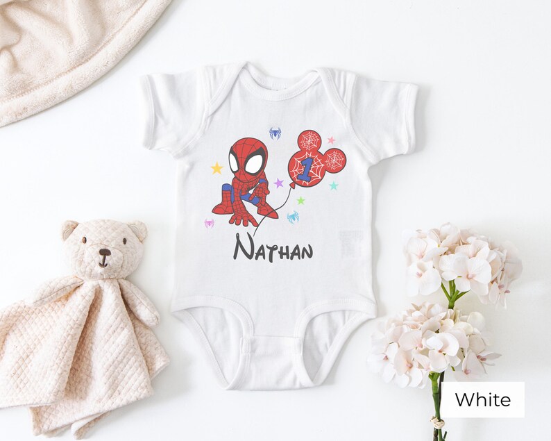 White baby onesie with custom Spiderman birthday design, personalized name Nathan and age 1, colorful stars, styled with teddy bear and flowers