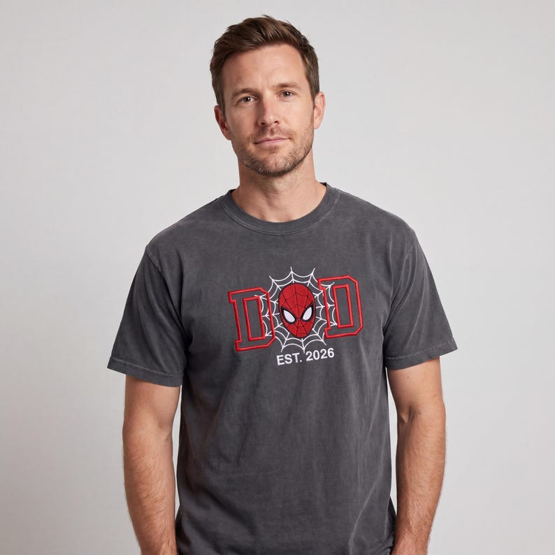 K&ouml;nnte beinhalten: Ein anthrazitfarbenes T-Shirt mit einem rot-wei&szlig;en Stickdesign. Das Design zeigt das Wort "DAD" mit einer Spider-Man-Maske in der Mitte und dem Text "EST. 2026" darunter. Das T-Shirt ist kurz&auml;rmlig.