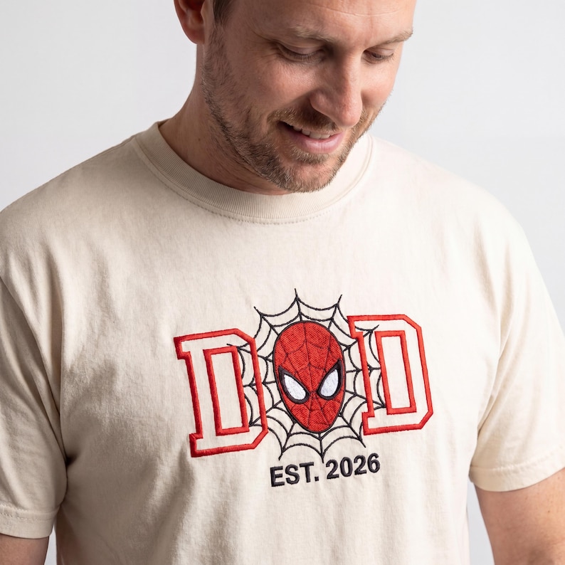 K&ouml;nnte beinhalten: Cremefarbenes T-Shirt mit dem Wort "DAD" in Rot, im Spider-Man-Stil. Die Spider-Man-Maske befindet sich in der Mitte, mit einem Spinnennetz-Design. Der Text "EST. 2026" steht darunter.