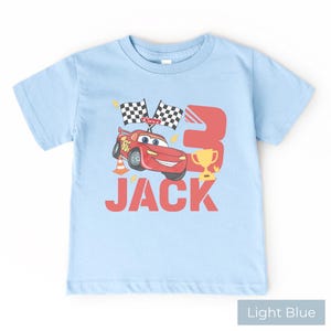 May include: A light blue short-sleeve t-shirt featuring a graphic of a red race car, two checkered flags, a traffic cone, a gold trophy, and the number 3. The name JACK is printed in large red letters below the car and trophy graphic.
