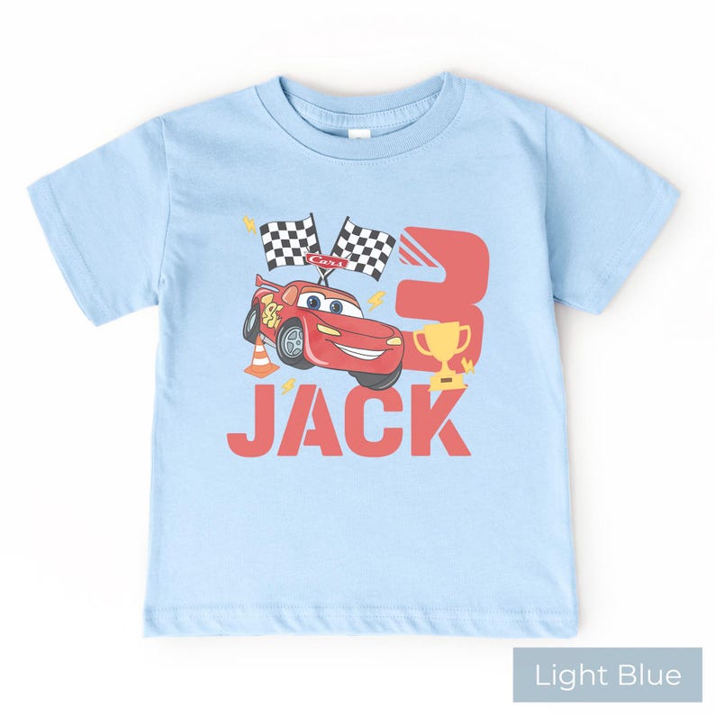 May include: A light blue short-sleeve t-shirt featuring a graphic of a red race car, two checkered flags, a traffic cone, a gold trophy, and the number 3. The name JACK is printed in large red letters below the car and trophy graphic.